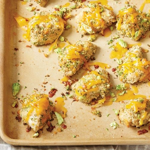 A stoneware baking sheet displays several pieces of broccoli-cheddar chicken nuggets. The nuggets are golden brown with a crispy breaded coating and are topped with melted cheddar cheese. Small bits of broccoli are mixed into the coating, adding a green hue to the nuggets. The baking sheet shows a few scattered pieces of broccoli and cheese, enhancing the appetizing appearance of the dish.