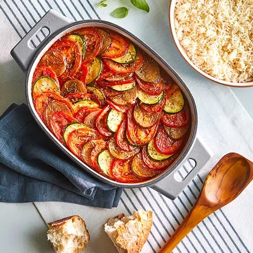 Colorful dish of Ratatouille, featuring a beautiful arrangement of thinly sliced vegetables like zucchini, yellow squash, eggplant, and tomatoes, layered in a circular pattern and baked to perfection. The vegetables are vibrant and glistening with a light coating of olive oil and herbs, showcasing a mix of textures and flavors in a rustic and appetizing presentation.