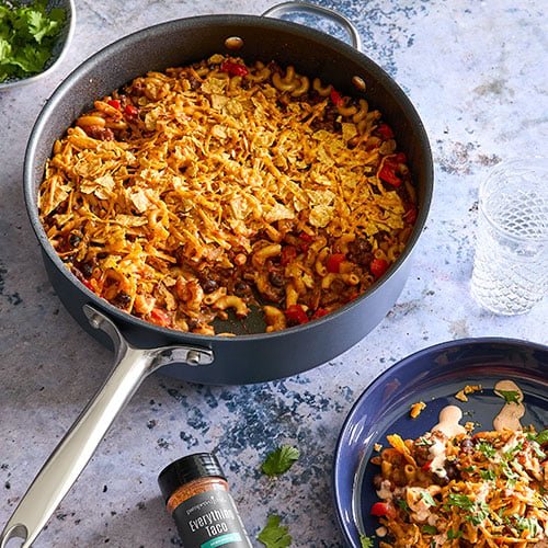 Skillet of taco chili mac, a hearty dish featuring macaroni pasta, ground meat, beans, and vegetables, all seasoned with taco spices and topped with melted cheese and crushed tortilla chips. A plate with a serving of the dish is placed beside the skillet, garnished with fresh cilantro. A bottle of 'Everything Taco' seasoning and a glass of water are also visible, adding to the inviting and flavorful meal setting.