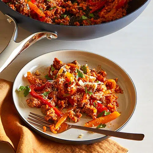 A plate of stuffed pepper skillet, featuring a hearty mixture of ground meat, rice, bell peppers, and tomato sauce, garnished with fresh parsley. The colorful dish is presented on a white plate with a fork, accompanied by an orange napkin. In the background, the skillet with the remaining stuffed pepper mixture is visible, highlighting the rich and flavorful ingredients.